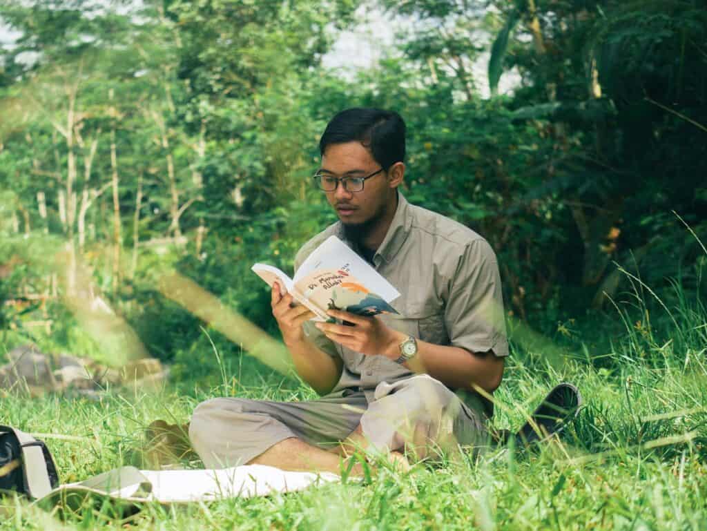 Young man sitting in the grass reading a book near a river, away from screens
