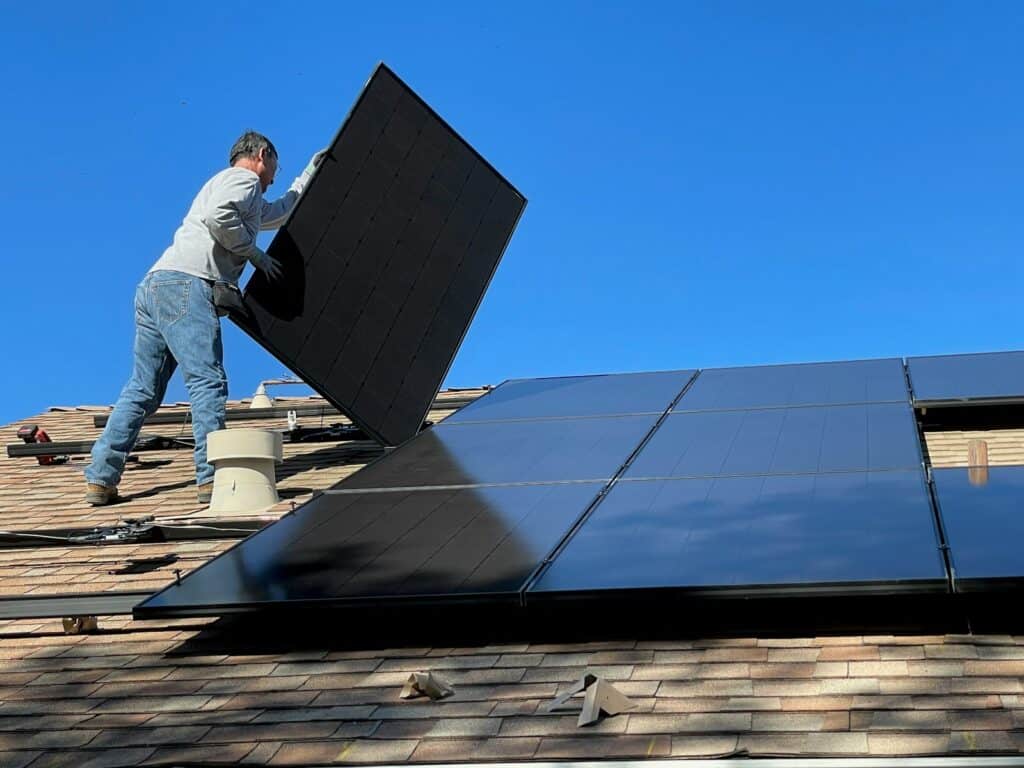 Worker fitting a black solar panel onto a shingled roof under a blue sky